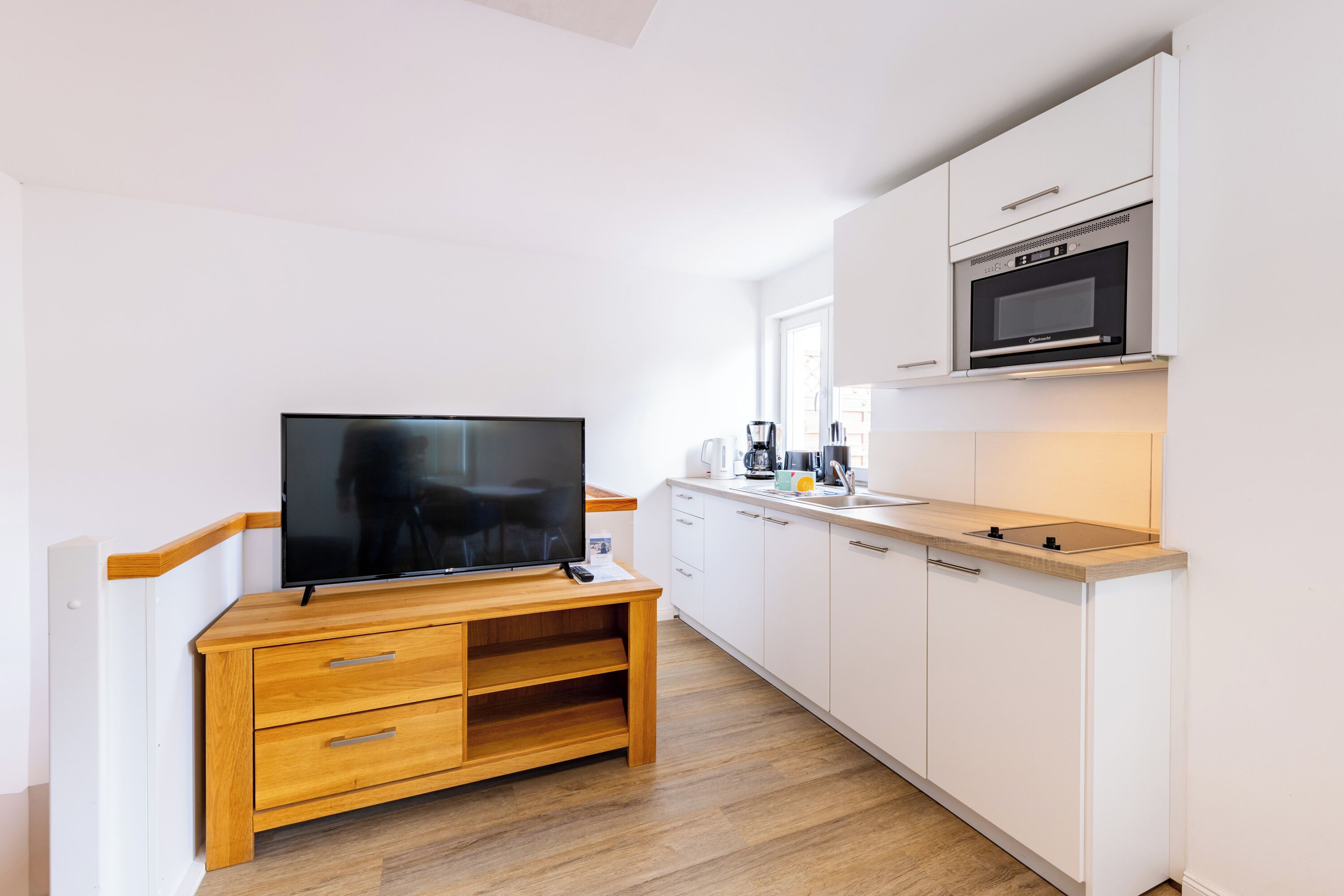 A compact kitchen with white cabinets, a microwave, a coffee maker, and a wooden entertainment unit with a flat-screen TV.