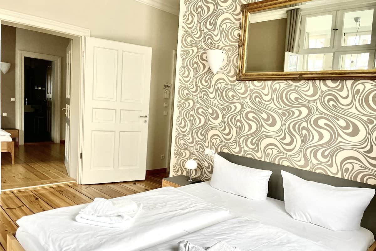 A hotel room with a wooden floor, a bed with white linens, a nightstand with a lamp, a mirror above the bed, and a patterned wallpaper.
