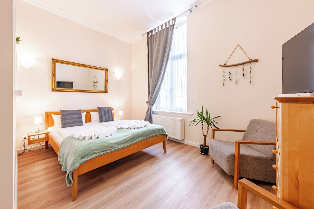 A bedroom with a wooden bed, a bedside table with a lamp, a chair, a TV, and a window with curtains.
