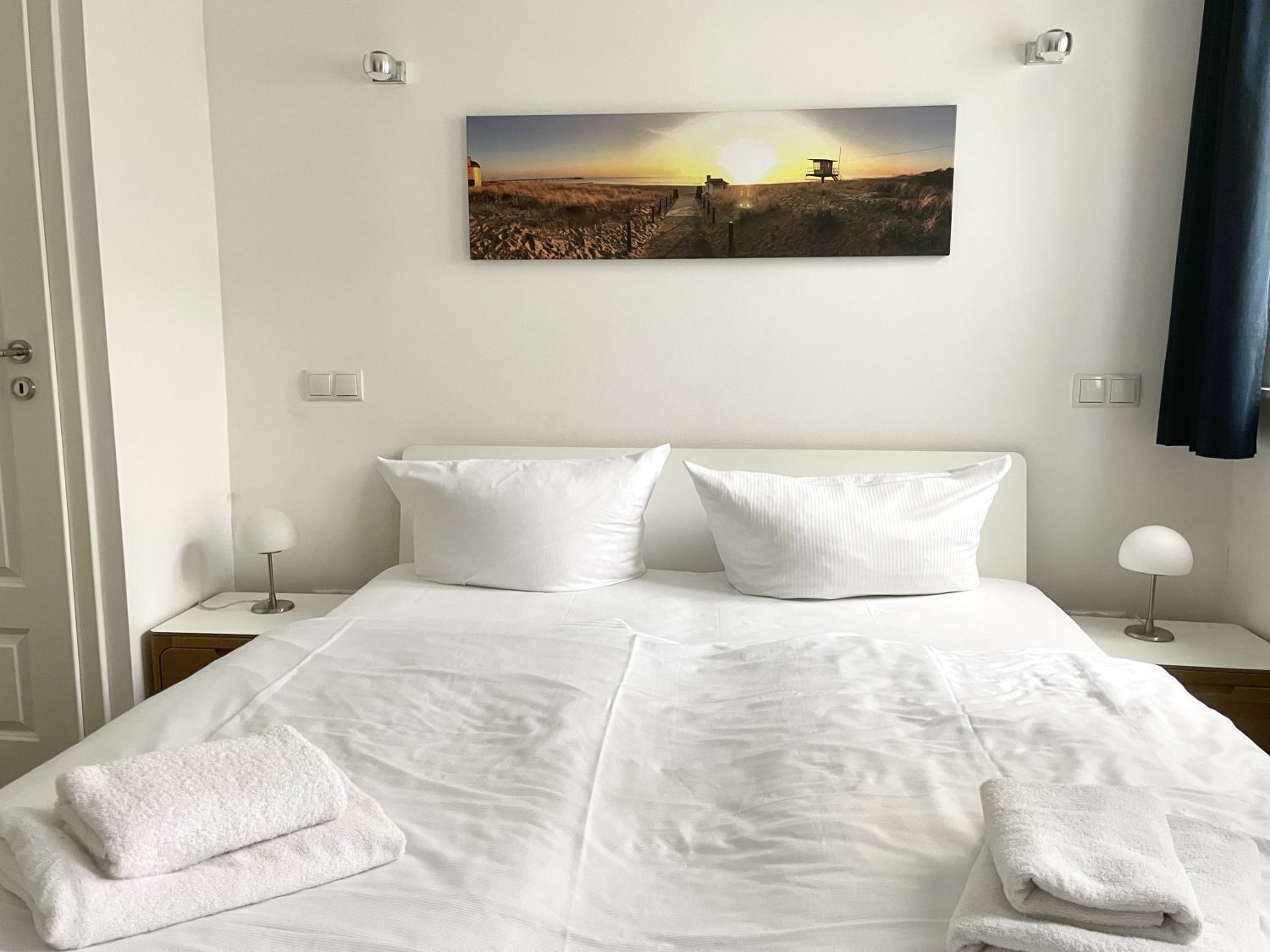 Una cama bien hecha con sábanas y toallas blancas, un cuadro colgado en la pared que muestra un atardecer sobre la playa, y dos lámparas de noche.