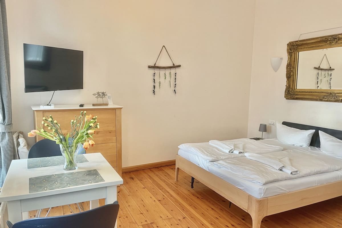A hotel room with a bed, a desk with a chair, a television, a wooden dresser, and a wall-mounted mirror.