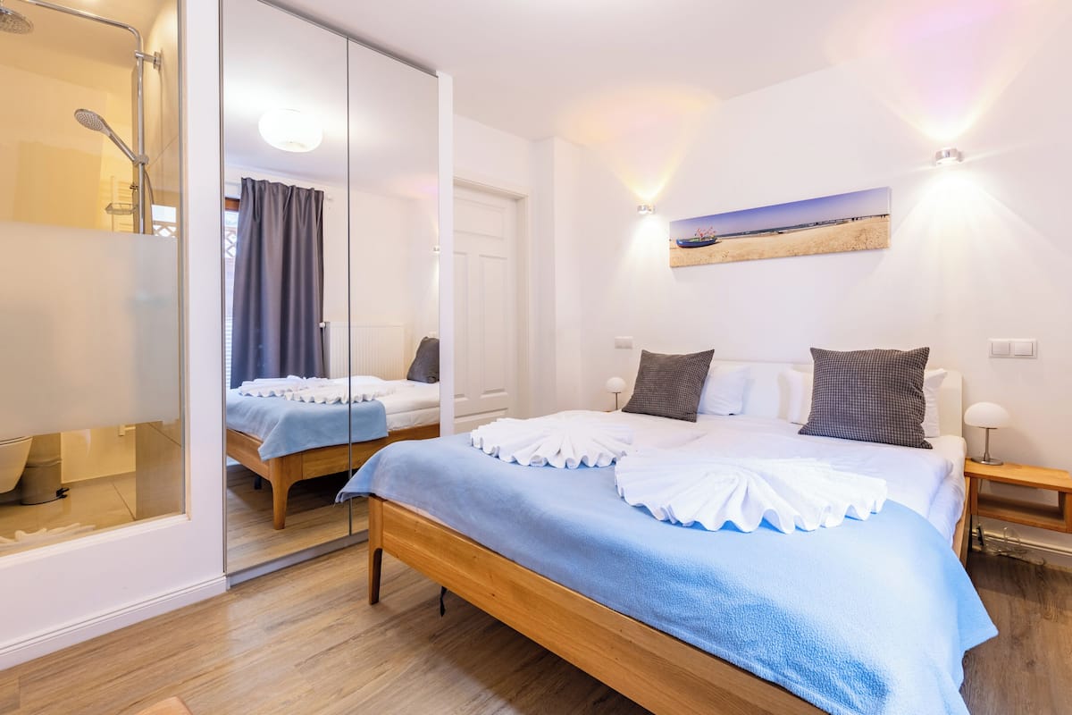 A modern bedroom with a large bed, a glass shower enclosure, and a view of the beach through a window.