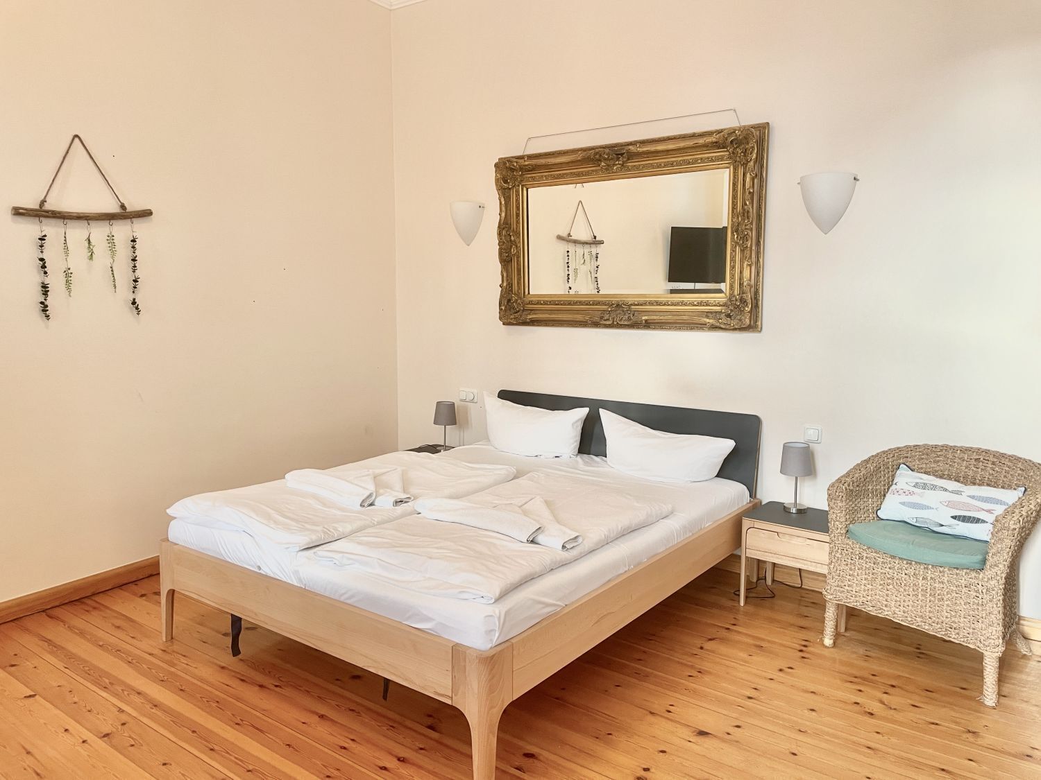 Een netjes opgemaakt bed met witte lakens, een houten hoofdbord en een nachtkastje met een lamp. Een rieten stoel met kussen staat naast het bed.