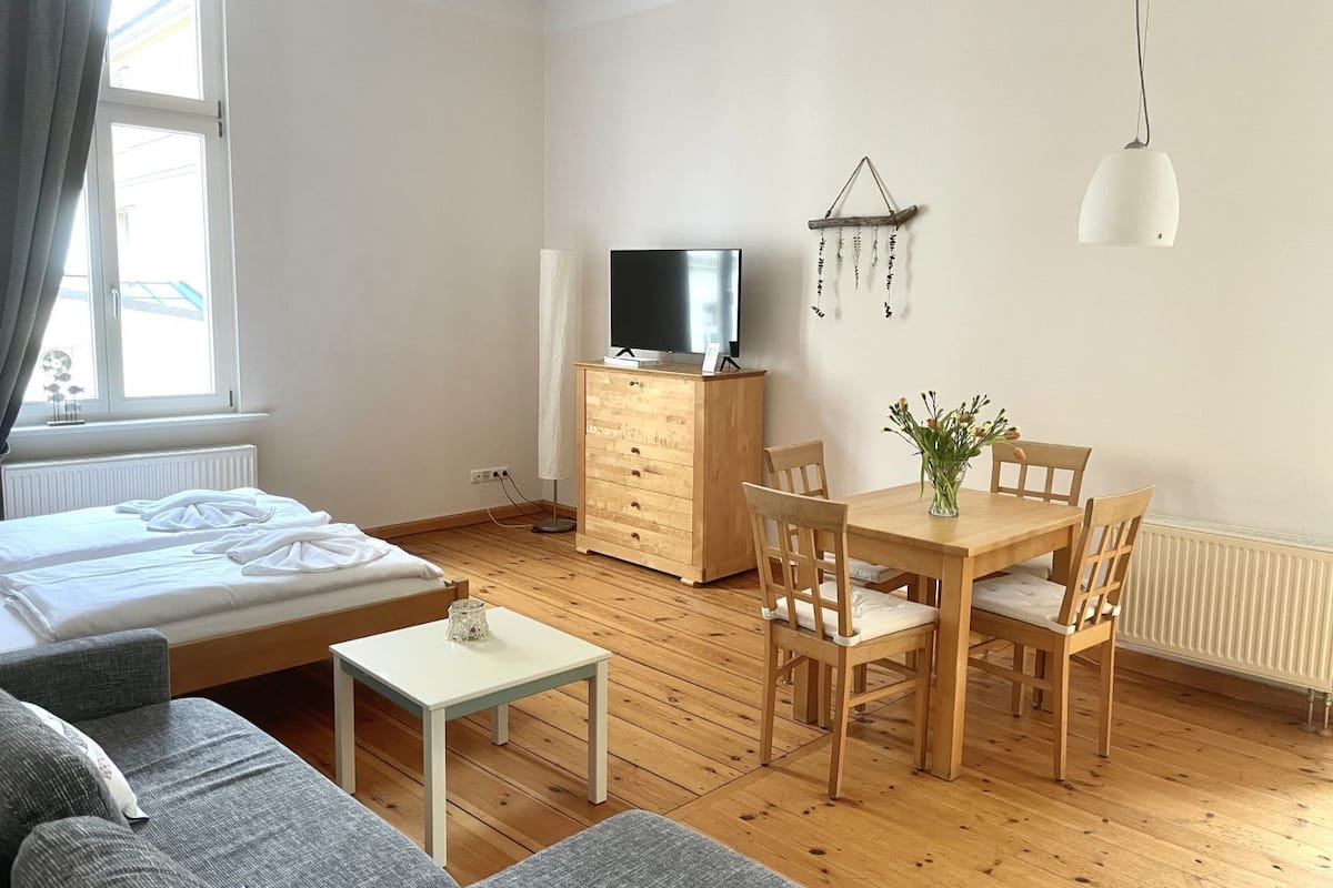 A modern, well-lit room with a bed, a sofa, a small table, a wooden cabinet, a TV, and a dining table with chairs.