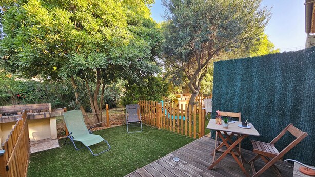 Une terrasse en bois avec une table et des chaises, entourée d’une clôture en bois et d’une haie verdoyante.