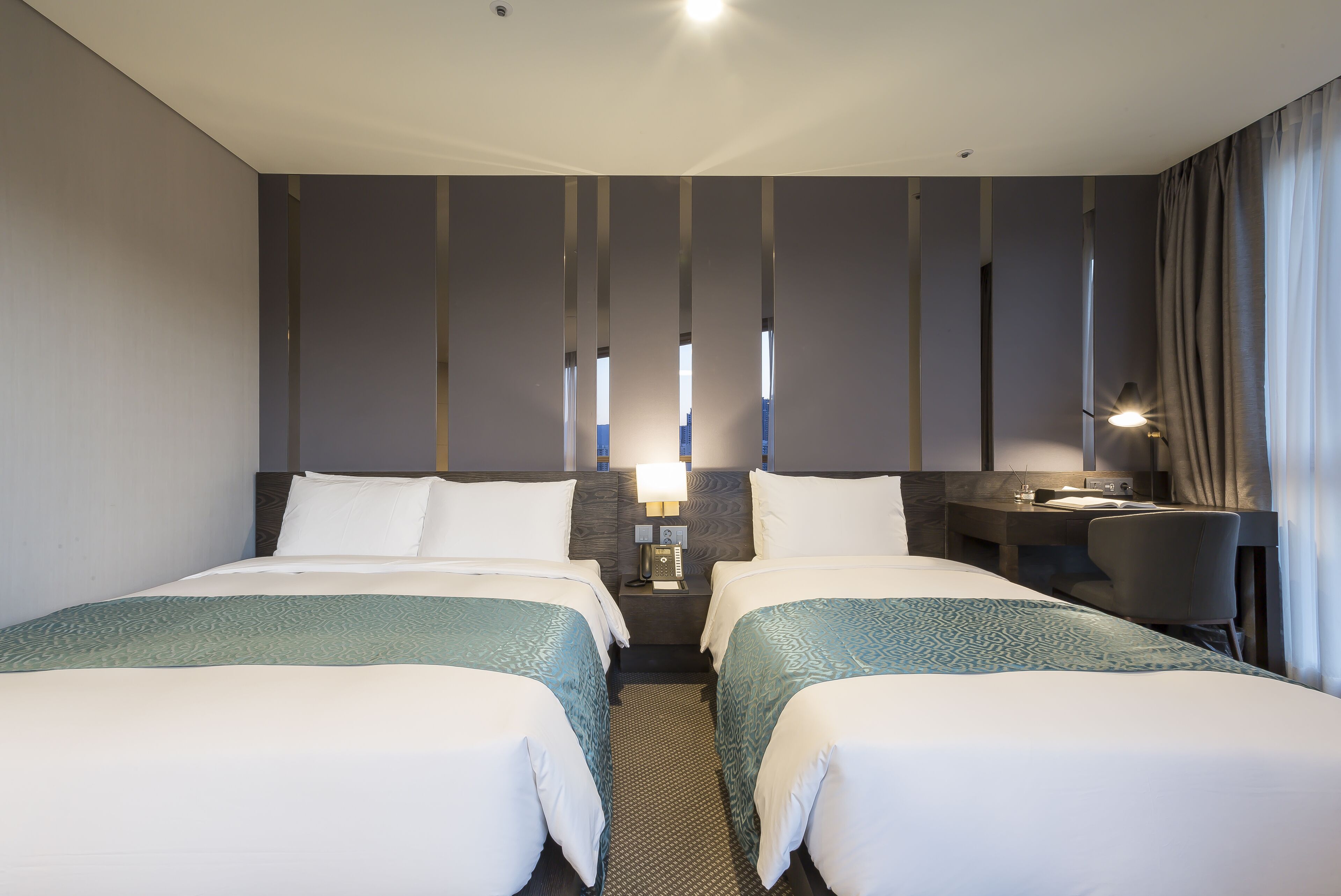 A hotel room with two beds, a desk, a chair, a nightstand with a lamp, and a window with curtains.