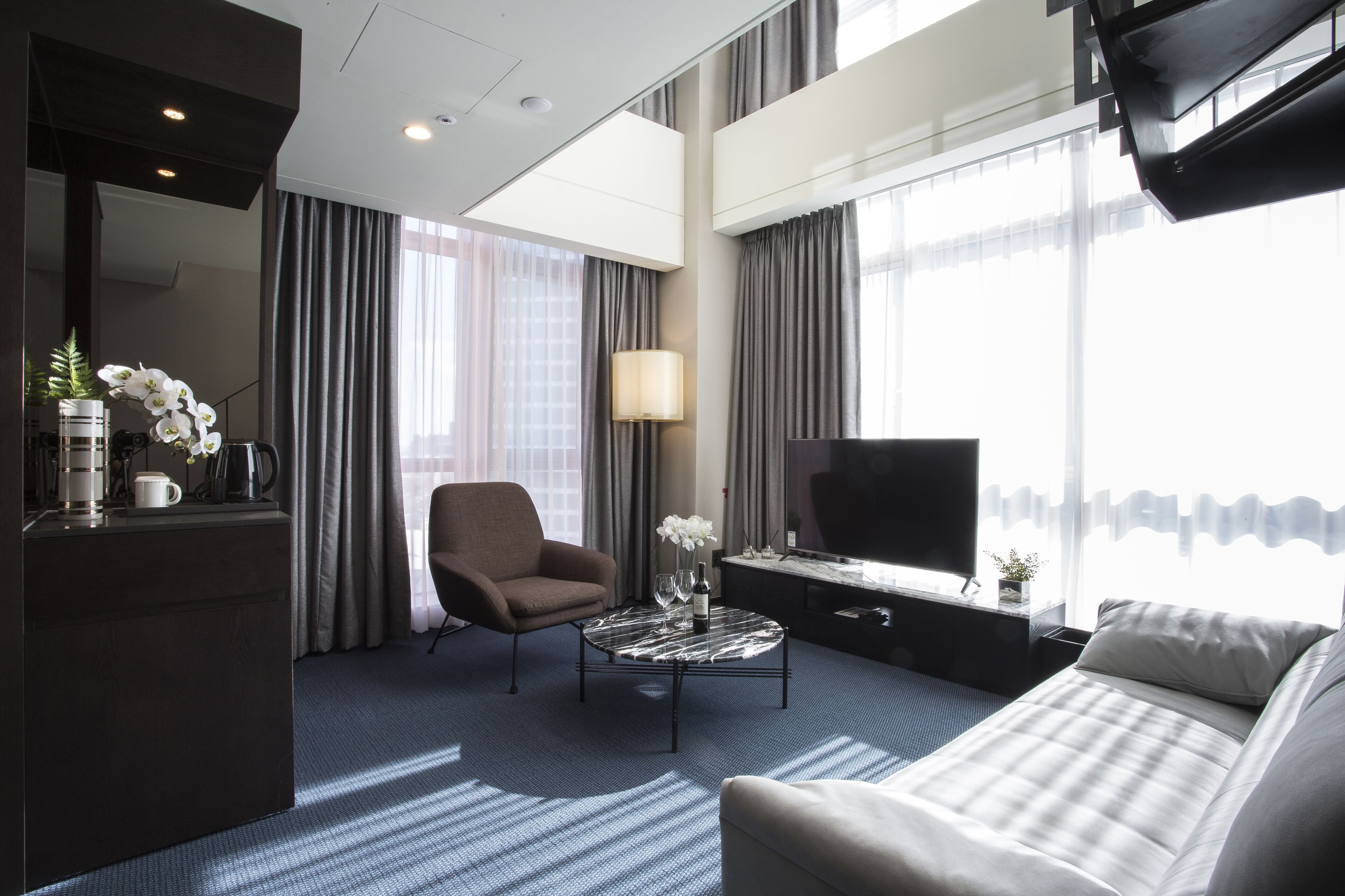 A modern hotel room with a sofa, armchair, coffee table, TV, and large windows with curtains.