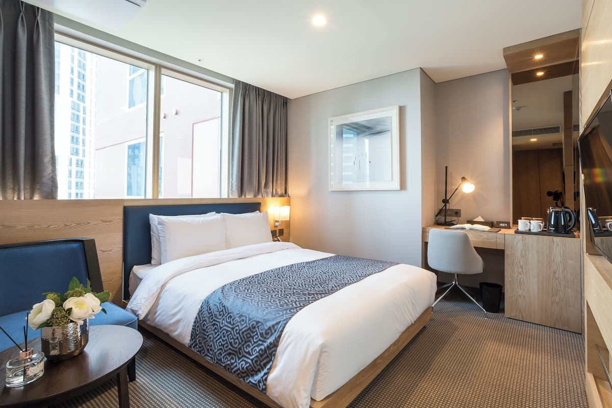 A hotel room with a bed, a desk, a chair, a small table with a vase of flowers, and a large window with curtains.