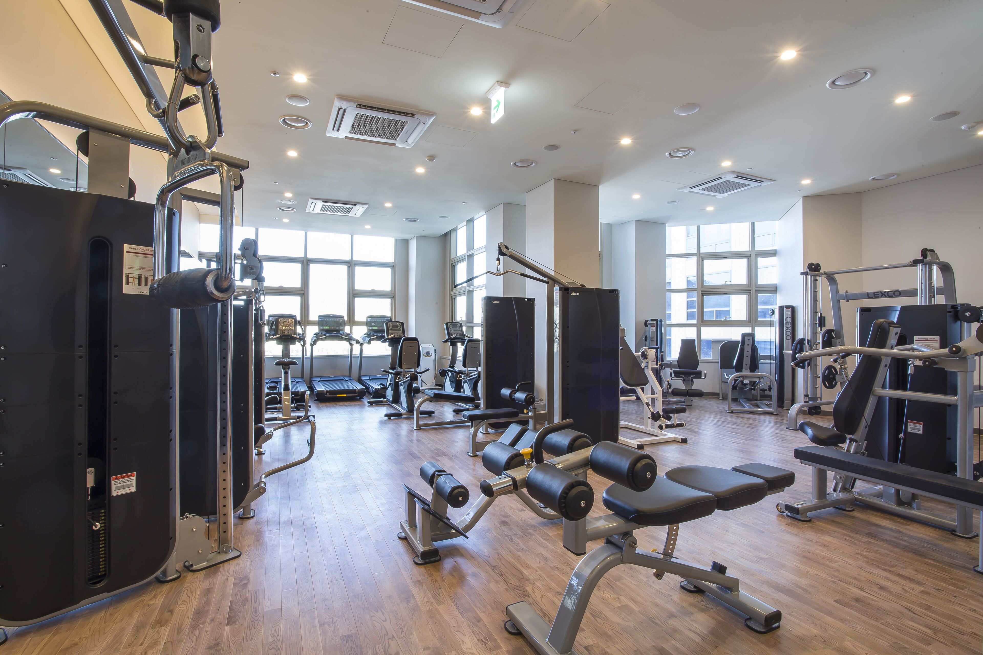 A gym with various exercise equipment, large windows, and a wooden floor.