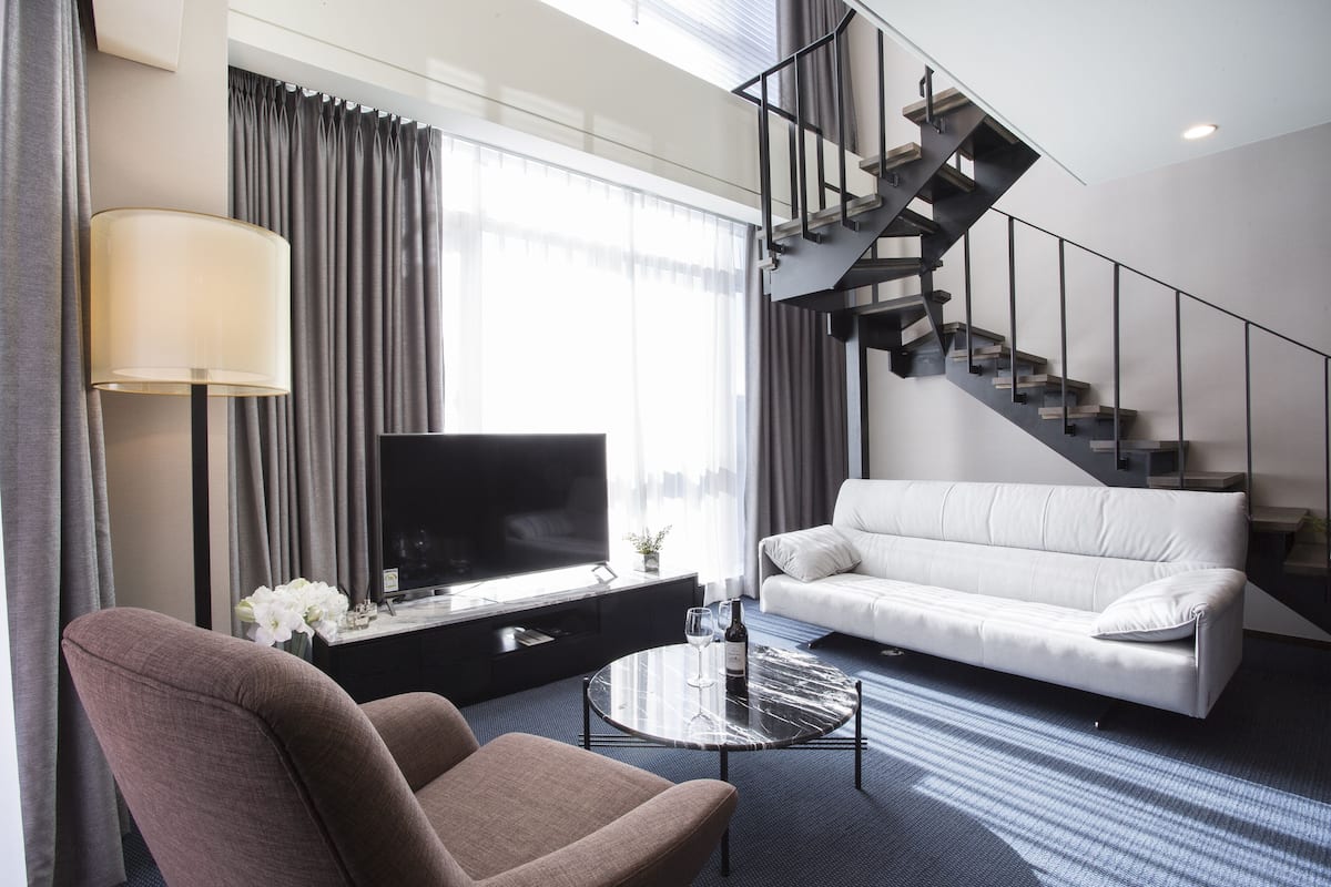 A modern hotel room with a sofa, armchair, flat-screen TV, and a staircase.