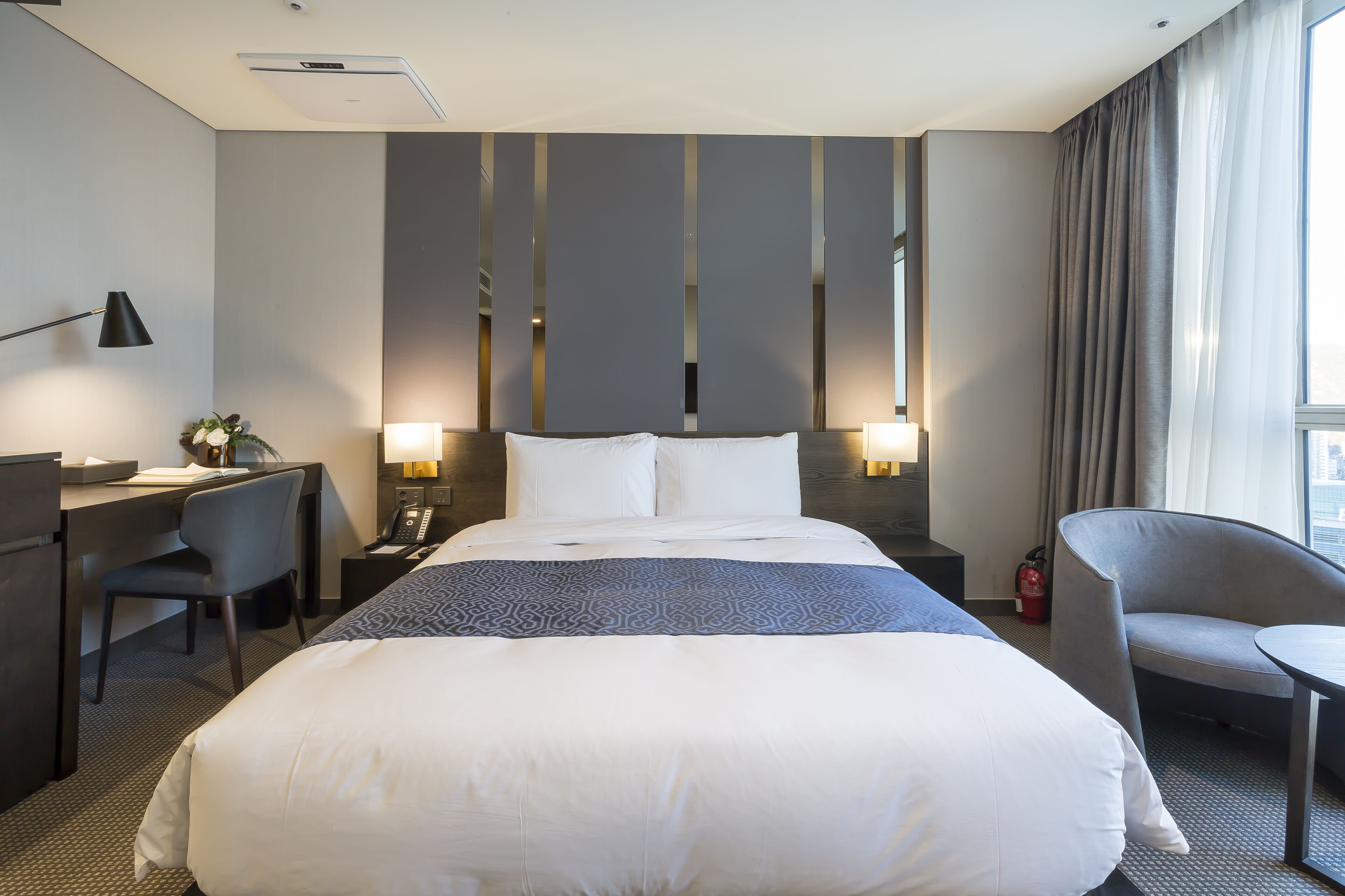 A hotel room with a large bed, a desk with a chair, a small table, and a chair. There are two bedside lamps, a fire extinguisher, and a window with curtains.