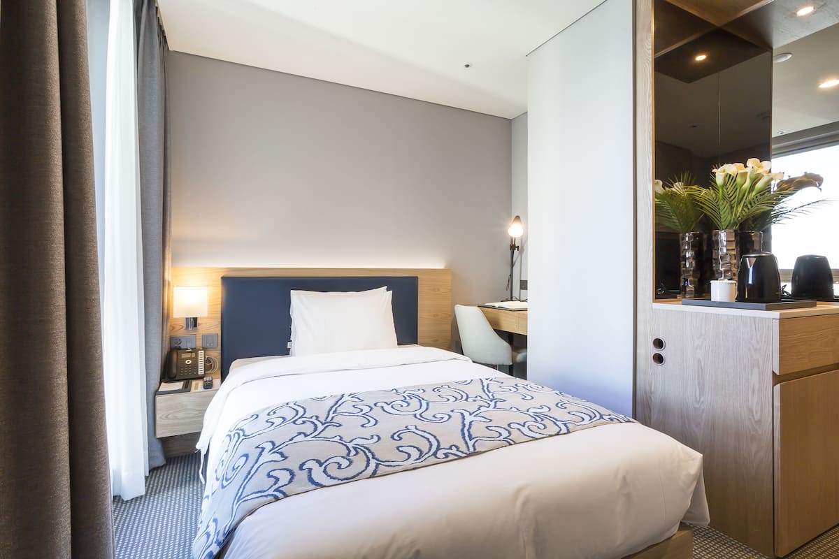 A hotel room with a bed, bedside table, lamp, and a small desk with a chair.