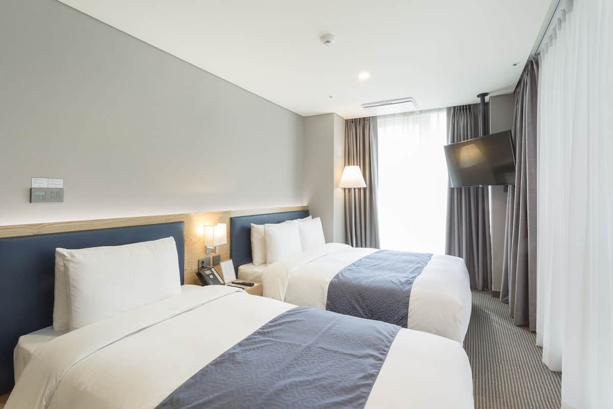 A hotel room with two beds, a TV, a lamp, and a window with curtains.