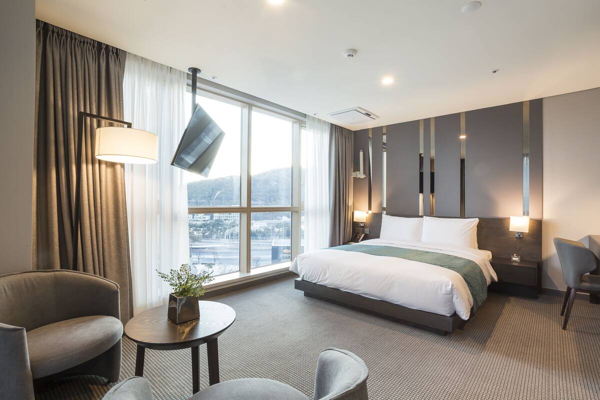 A modern hotel room with a large bed, a sitting area with a table and chairs, a TV mounted on the wall, and a view of the city through large windows.