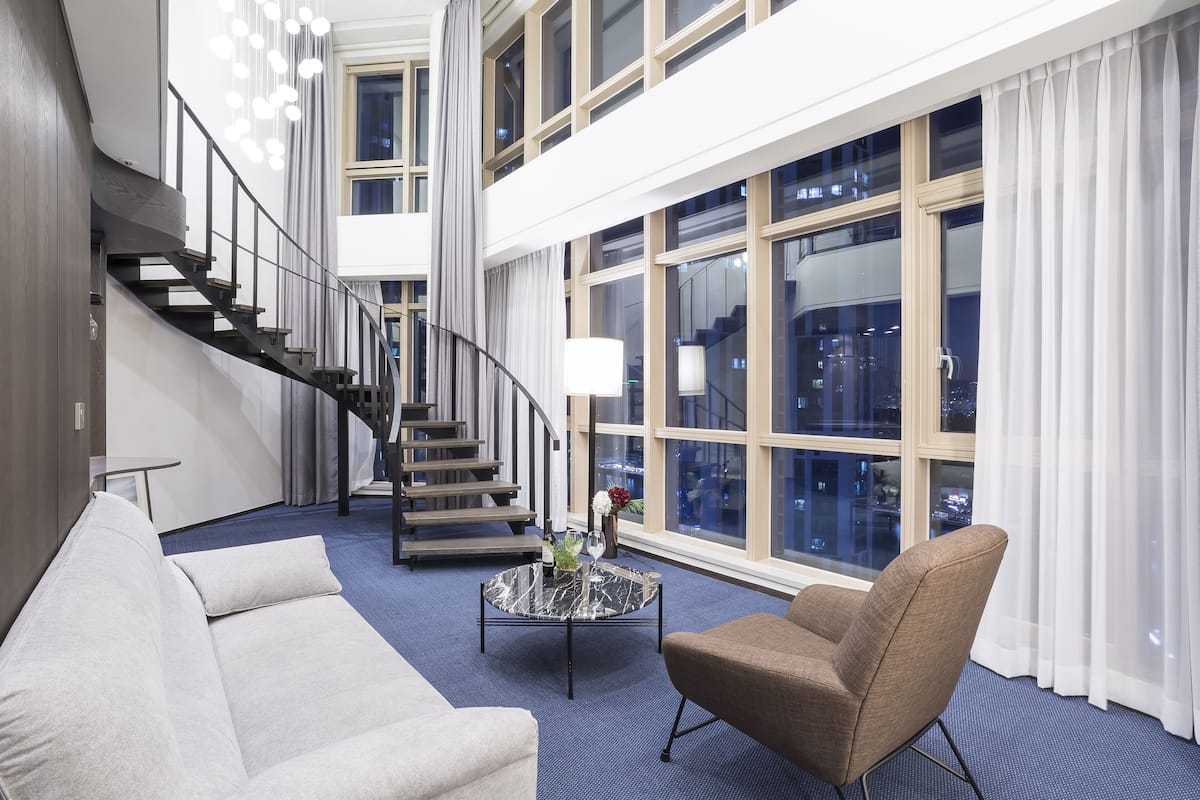 A modern hotel lobby with a curved staircase, a round marble coffee table, a beige armchair, and large windows offering city views.