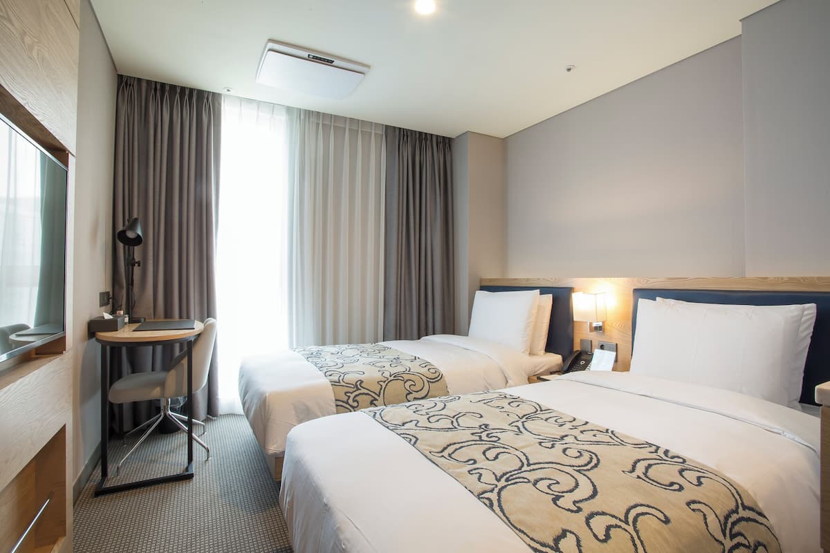 A hotel room with two beds, a desk, a chair, and a window with curtains.