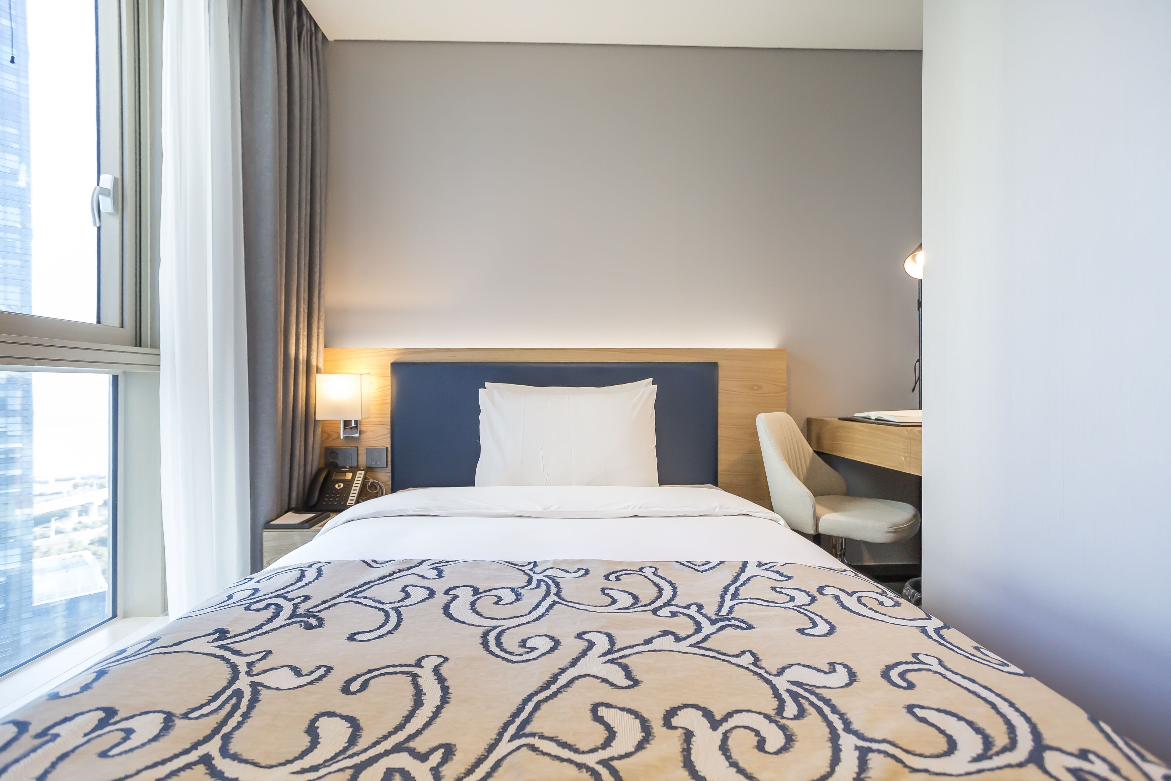 Una habitación de hotel con cama, escritorio, silla, lámpara y ventana con cortinas.