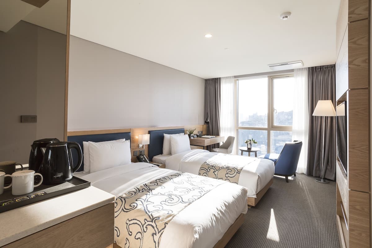 A hotel room with two beds, a desk, a chair, a coffee maker, and a view of the city.