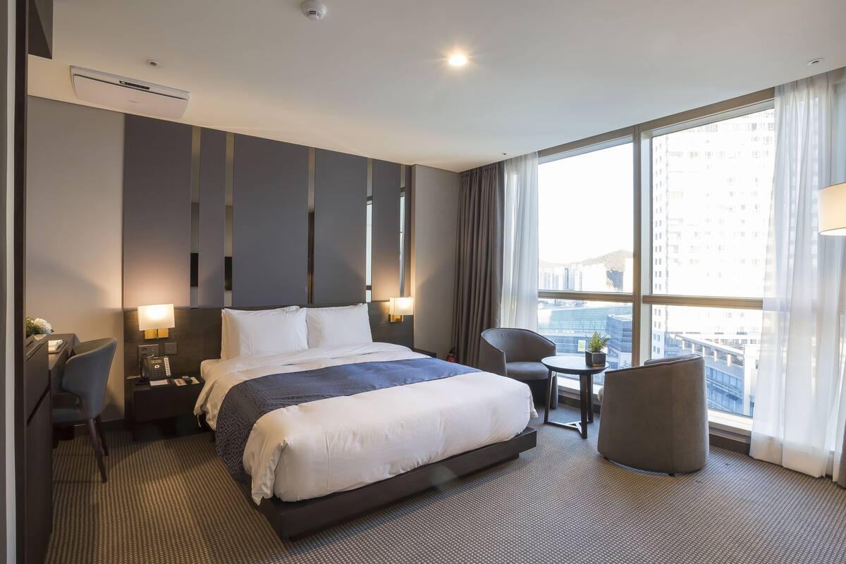 A modern hotel room with a large bed, a desk, a chair, and a view of the city through large windows.
