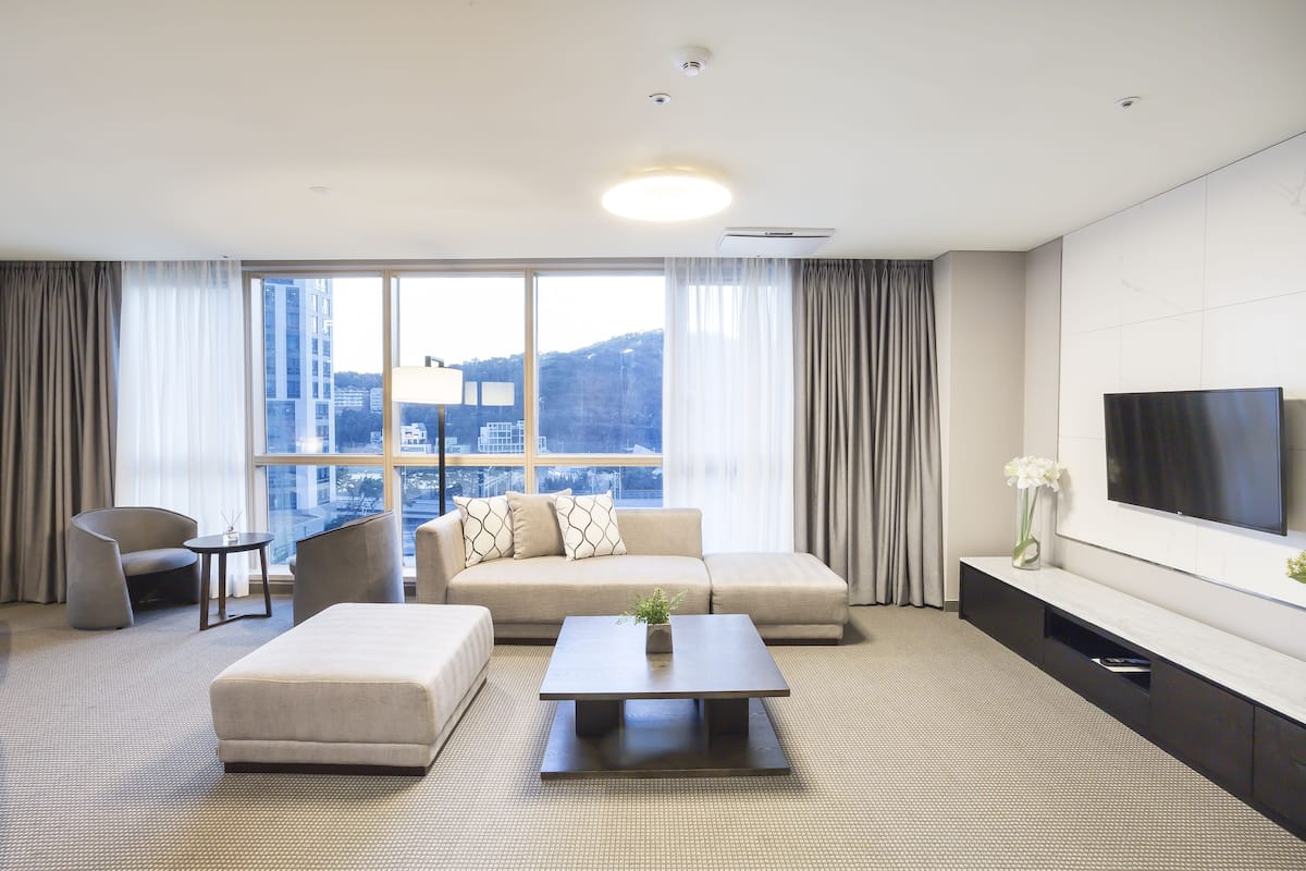 A modern hotel room with a large window, a sofa, a coffee table, a TV, and a small side table.