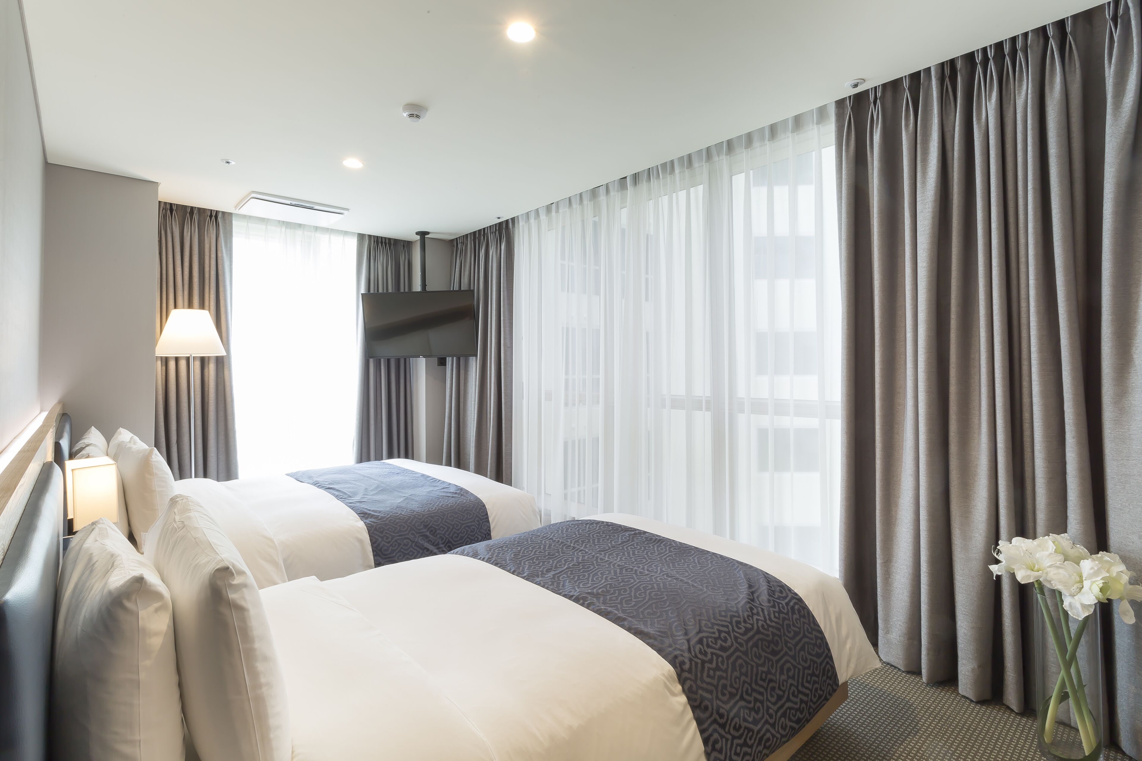 A hotel room with two beds, a flat-screen TV, a lamp, and a vase with flowers.