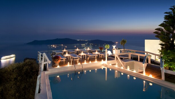 A poolside area with lounge chairs and tables, a staircase leading down to the pool, and a view of the sea at dusk. - Artemis Villas (Santorini)