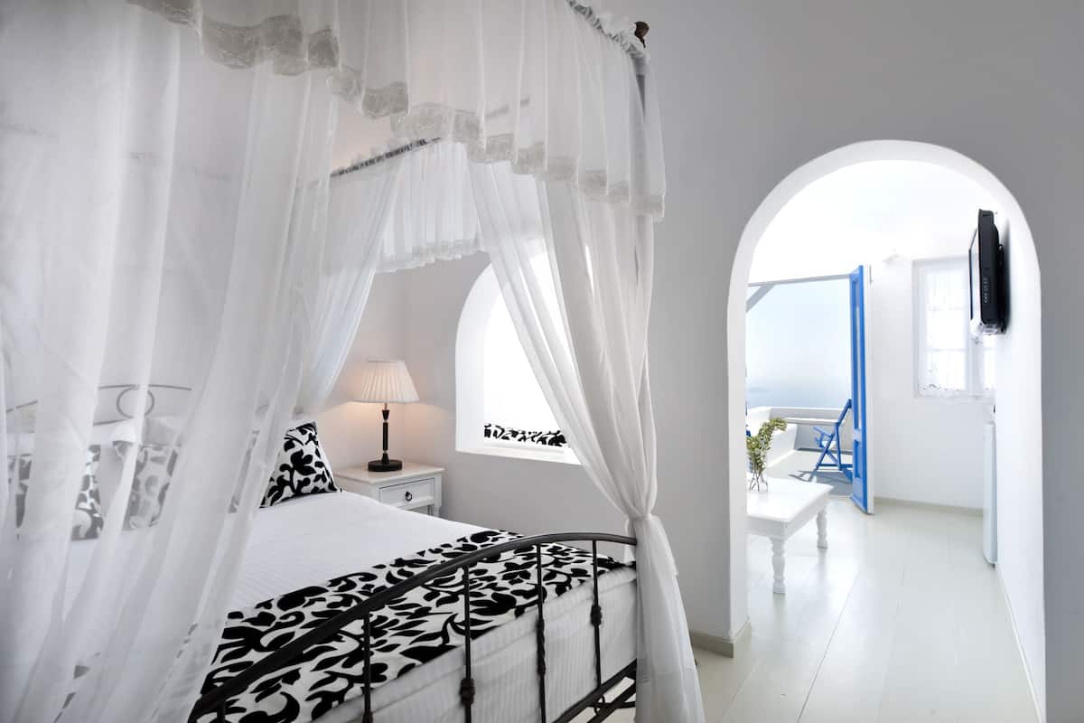 A bedroom with a canopy bed, a bedside table with a lamp, and a view of the outside through an arched doorway.