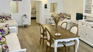 A dining area with a wooden table and chairs, a white sofa, and a cabinet with a television. - Artemis Villas (Santorini)