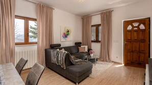 Una sala moderna con un sofá gris, una mesa de centro y un comedor con sillas. Hay una puerta de madera y una ventana con cortinas.