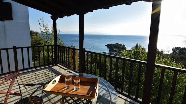 Ein Balkon mit Holztisch und -stühlen, einem Tablett mit Gläsern und Blick auf das Meer und die grüne Landschaft.