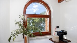 A room with a white wall, a wooden-framed arched window, a potted plant, a coffee maker, and a small decorative item on the counter.