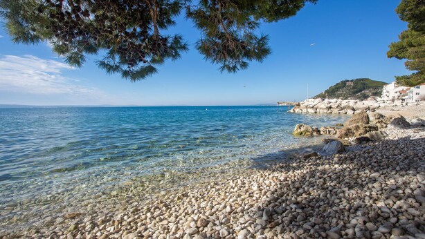 A pebble beach with clear water, a rocky shoreline, and a distant view of a coastal town.