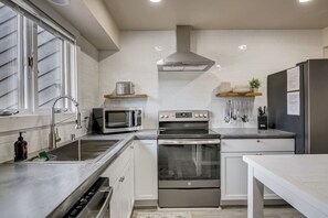 Una cocina moderna con electrodomésticos de acero inoxidable, armarios blancos y un ventanal con persianas.