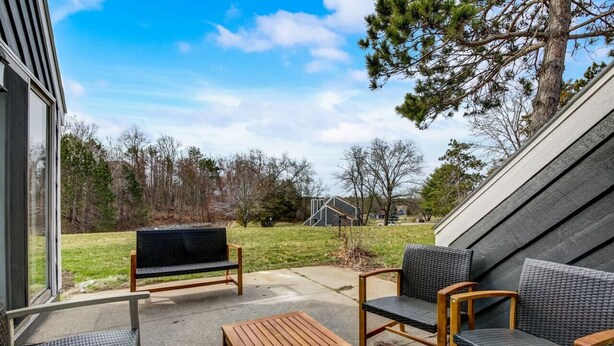 An outdoor patio with a wooden table and two wicker chairs, a bench, and a view of a grassy yard and trees.
