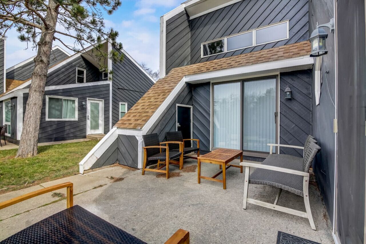 Un edificio moderno de dos pisos con techo inclinado, un patio cubierto con mobiliario exterior y un amplio ventanal corredizo.