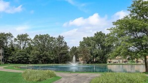 A serene park with a fountain, a pond, and a pathway leading to a building.