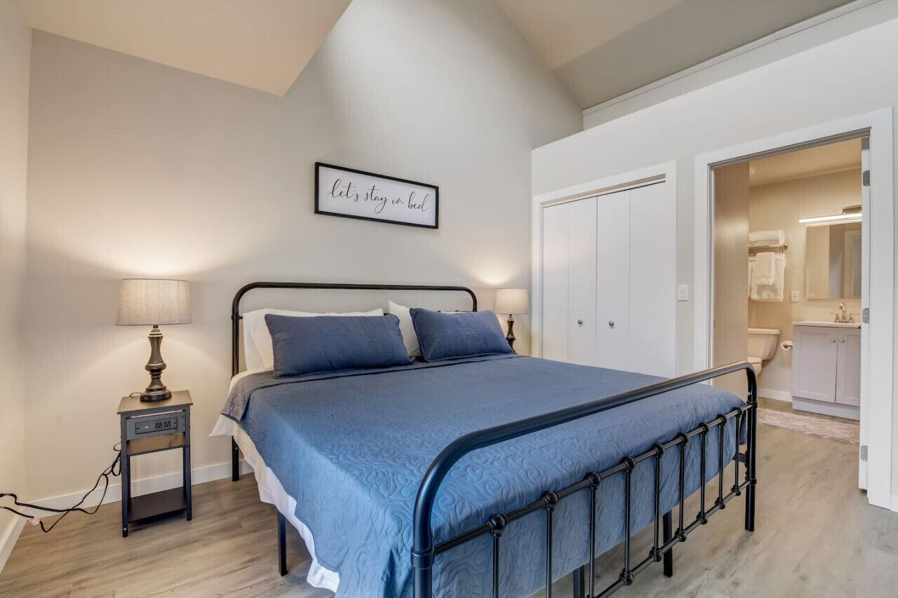 Une chambre à coucher comprenant un lit, des lampes de chevet, un panneau décoratif fixé au mur et une porte donnant accès à une salle de bain.