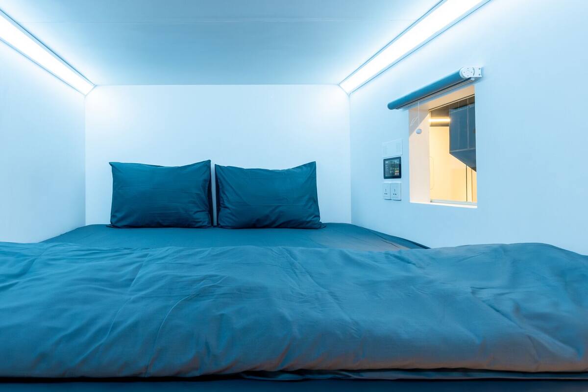 Ein kleiner, moderner Raum mit einem Bett, zwei Kissen und einem Fenster mit Jalousien.