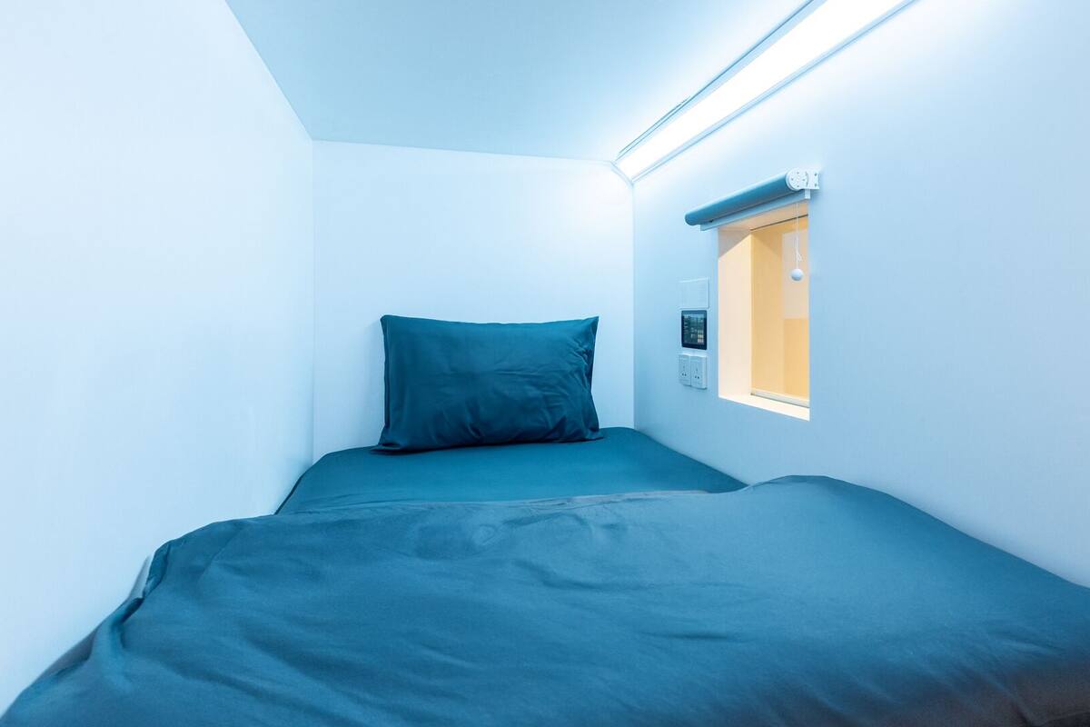Ein kleines Zimmer mit einem Einzelbett, einem Fenster, einem Wandthermostat und einer Deckenleuchte.