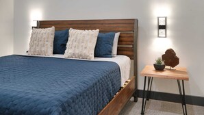 A bed with a wooden headboard, a blue quilted bedspread, and two pillows. There is a small wooden nightstand with a plant and a decorative object. - Vizcaya #2 - Moab's Newest Luxury Rental (Hot Tub) (Moab)