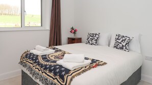 A bedroom with a large bed, a window with a view of a green field, a wooden nightstand with a vase of flowers, and patterned pillows. - Narracott Down (South Molton)