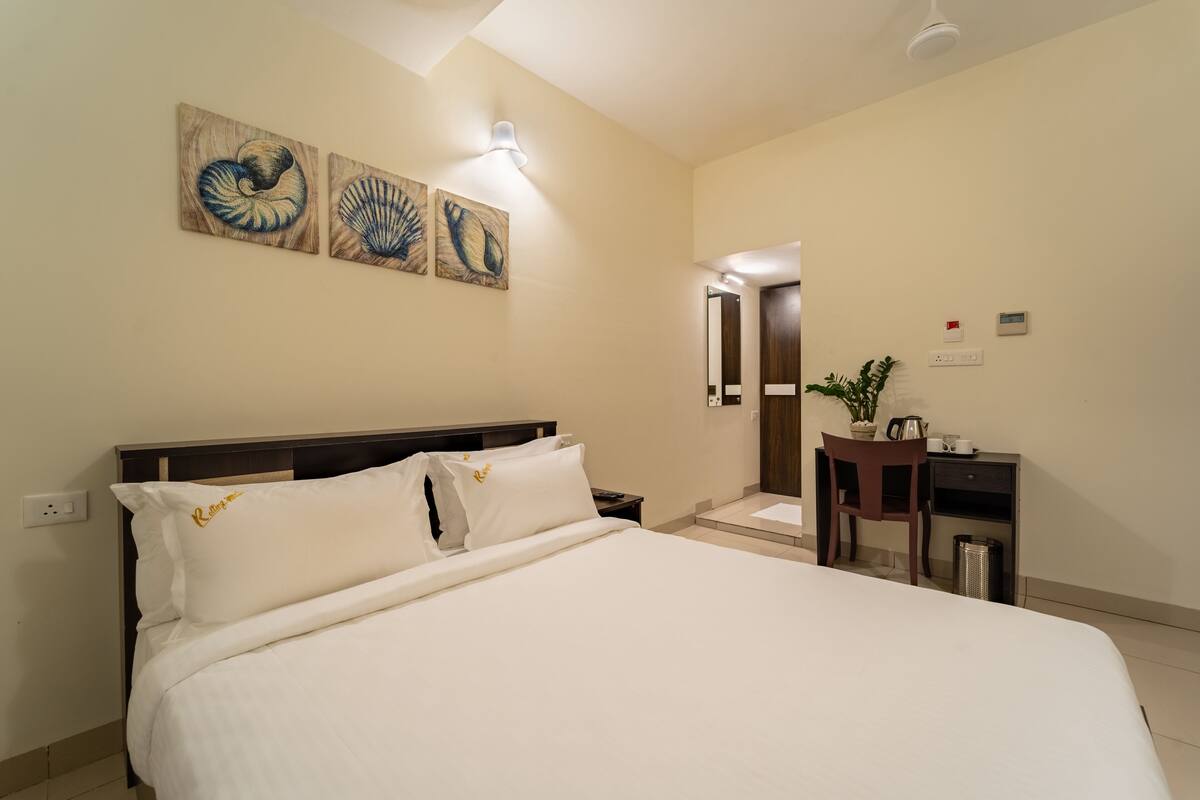 A hotel room with a large bed, a desk with a chair, a potted plant, and artwork on the wall.