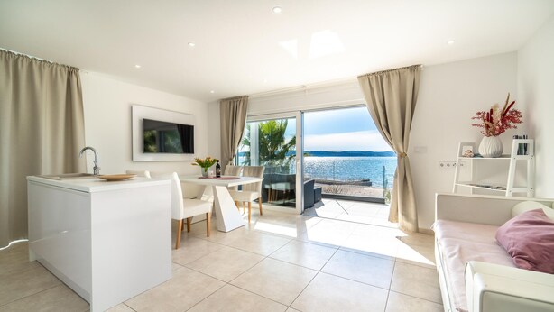 Een moderne, goed verlichte woonkamer met een keuken, een eettafel en een grote schuifpui naar een balkon met uitzicht op zee.
