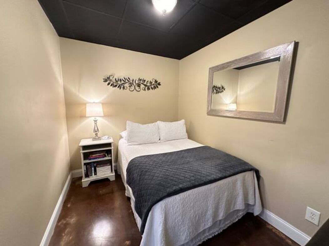 A bedroom with a bed, a nightstand with a lamp, a bookshelf, and a mirror on the wall.