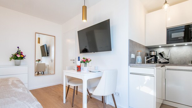 Una habitación de hotel moderna con cama, un área de comedor con mesa y sillas, una cocina con microondas y cafetera, y un televisor de pantalla plana grande montado en la pared.