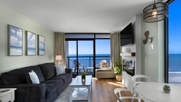 Ein modernes Wohnzimmer mit einem großen Fenster mit Blick auf den Ozean, einem schwarzen Sofa, einem weißen Couchtisch und einem Essbereich mit weißen Stühlen.