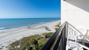 Ein Balkon mit weißen Plastikstühlen, der auf einen Strand mit sanften Wellen und einem klaren Himmel blickt.