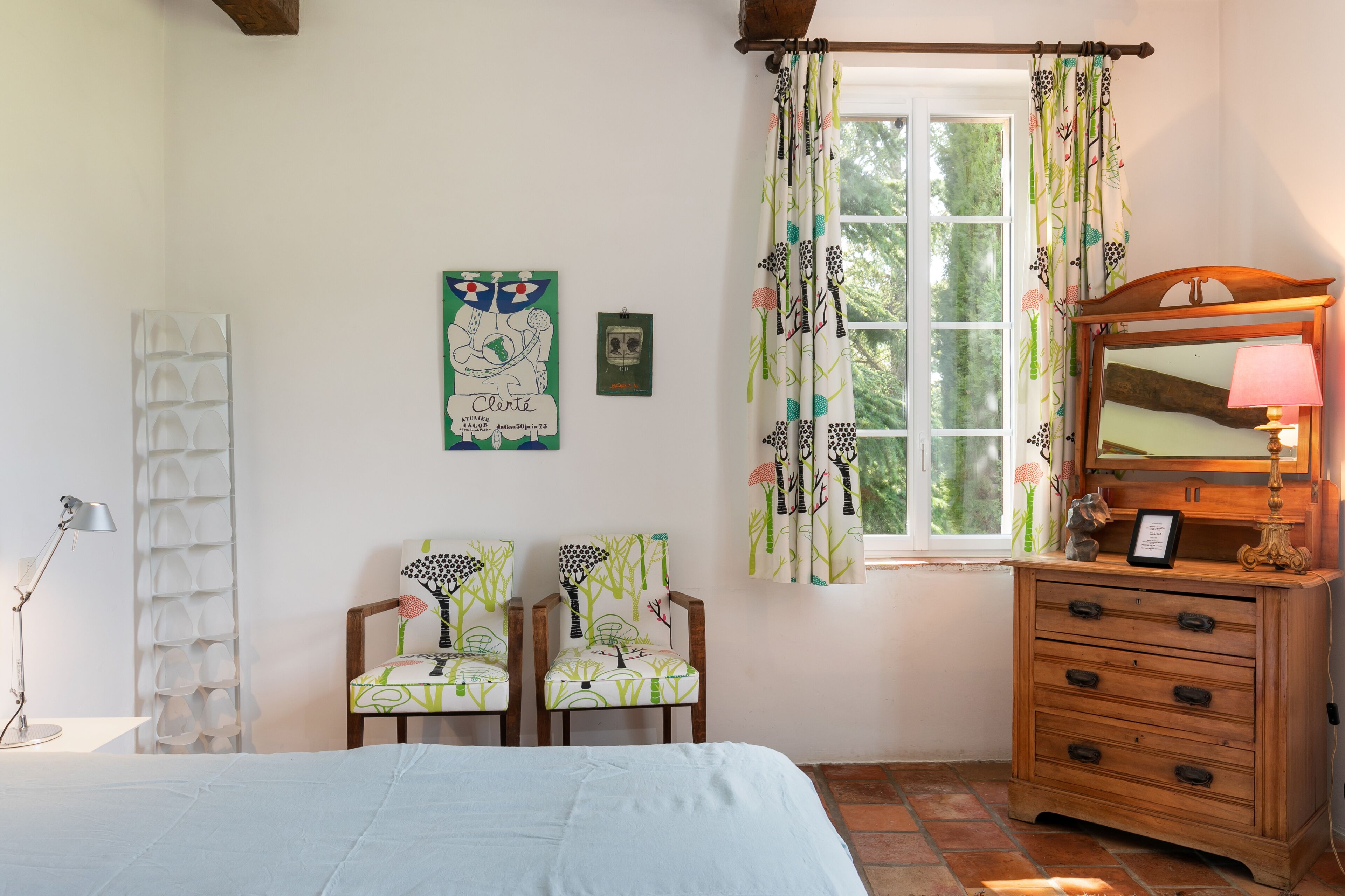 Een slaapkamer met een bed, twee stoelen, een dressoir, een spiegel en een raam met gordijnen.