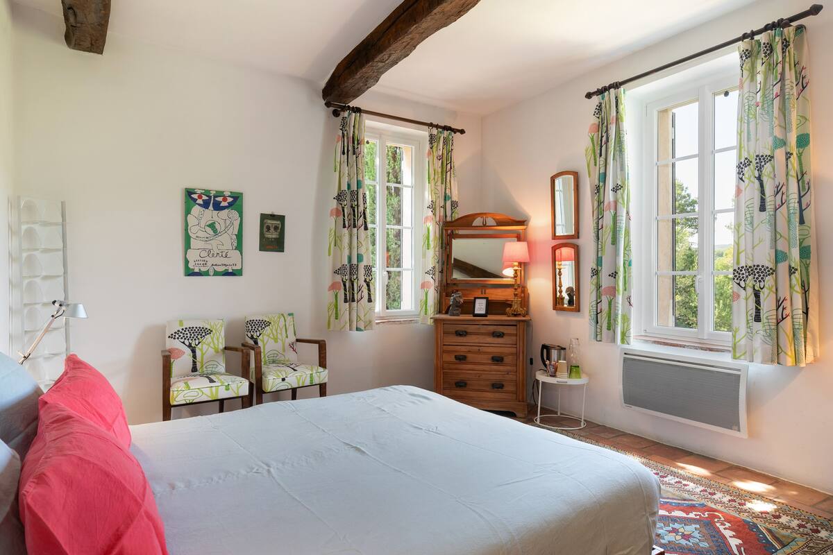 Ein Schlafzimmer mit einem Bett, zwei Stühlen, einer Kommode, einem Spiegel und einem Fenster mit Vorhängen.