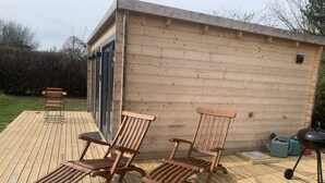 Una terraza de madera con dos sillones y una mesita, una cabaña de madera con una puerta, y equipo para cocinar al aire libre.